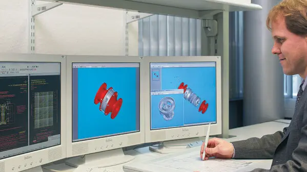 An engineer uses a stylus at a desk while three monitors display exploded-view 3D CAD models of a red mechanical component.