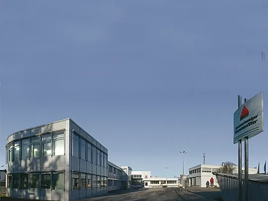 Modern industrial building complex with glass-fronted offices and a company sign at the entrance under a clear sky.