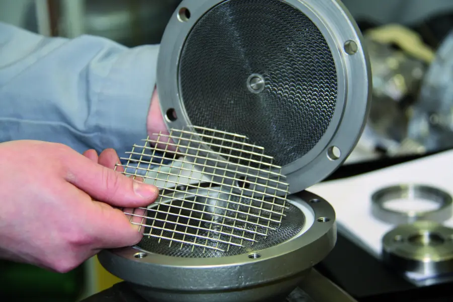 Close-up of hands holding a square metal mesh screen in front of an open circular metal filter housing in a workshop.