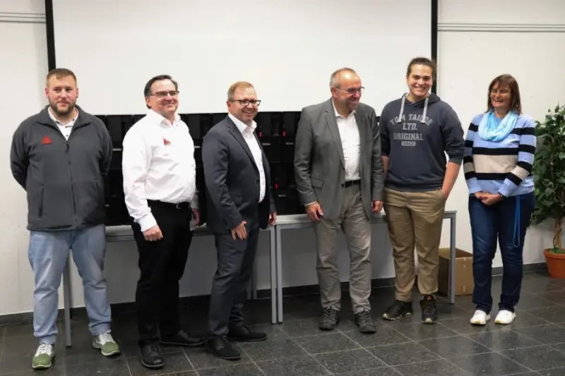 Six people stand smiling for a group photo in a classroom, in front of a table lined with black equipment cases and a projector screen.