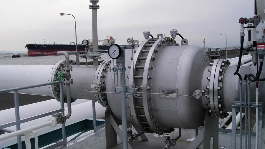 An industrial pipeline with a large bolted pressure vessel and gauges sits on a waterfront platform, with a cargo ship in the background.