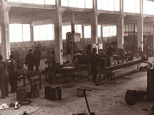 Vintage sepia-toned photo of a spacious factory workshop with workers standing at long tables and heavy machinery beneath tall windows and concrete columns.