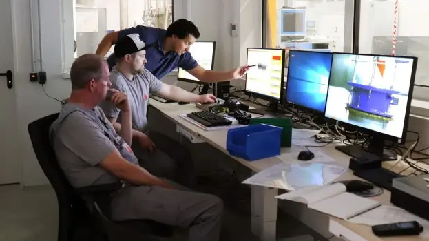 Three engineers collaborate at a control desk, reviewing test data and images on multiple computer monitors inside a technical laboratory.