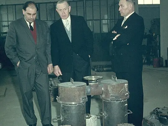 Three formally dressed men in suits stand in an industrial workshop, closely examining several cylindrical metal devices placed on the floor in front of them.